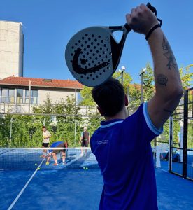 Ein Padelspieler im blauen Sechzig Trikot hebt den Arm zum Schlag, auf der anderen Seite des Netzes stehen einige andere Personen, die gerade Bälle aufheben.
