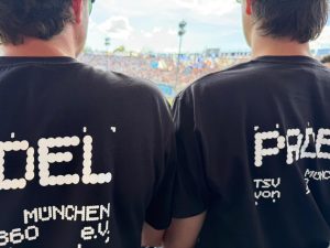 Zwei Mitglieder der Padel Sparte im Member-only T-Shirt der Sparte. Beschriftet ist das schwarze Shirt auf dem Rücken in groß mit dem Wort "Padel", bei welchem die weißen Buchstaben aus Padelbällen bestehen.Darunter steht TSV München von 1860 e.V über zwei Zeilen in kleineren Buchstaben.